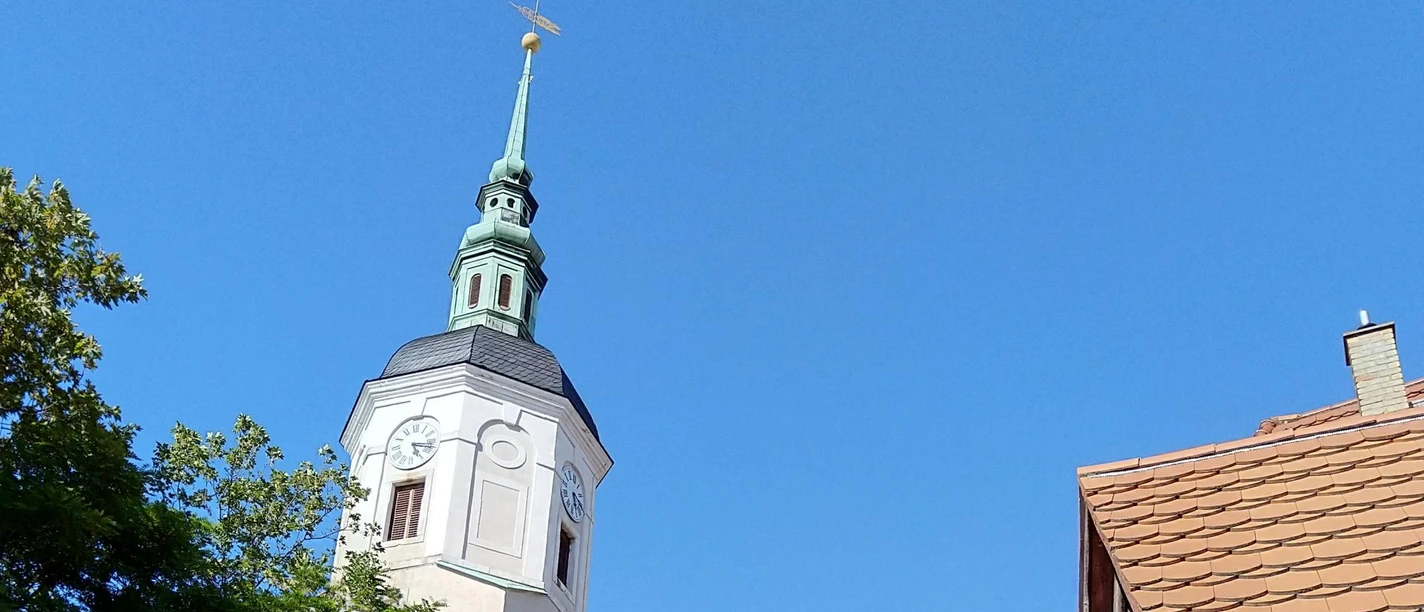 De kerktoren met zijn groene dak en windwijzer steekt boven de bomen en geplaveide straat uit, geflankeerd door oude gebouwen onder een strakblauwe hemel.