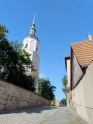 Stadtkirche Dohna The church tower with its green roof and weather vane rises above the trees and cobbled street, flanked by old buildings, under a clear blue sky.