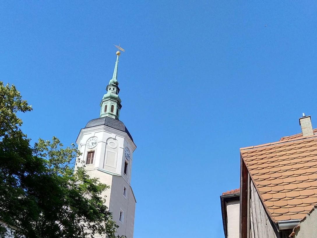Stadtkirche Dohna Kirchturm mit grünem Dach und Wetterfahne ragt über Bäume und Kopfsteinpflasterstraße, flankiert von alten Gebäuden, unter klarem, blauem Himmel.The church tower with its green roof and weather vane rises above the trees and cobbled street, flanked by old buildings, under a clear blue sky.Kostelní věž se zelenou střechou a větrolamem se tyčí nad stromy a dlážděnou ulicí, lemovanou starými budovami pod jasně modrou oblohou.Wieża kościoła z zielonym dachem i wiatrowskazem wznosi się ponad drzewami i brukowaną ulicą, otoczona starymi budynkami pod czystym, błękitnym niebem.De kerktoren met zijn groene dak en windwijzer steekt boven de bomen en geplaveide straat uit, geflankeerd door oude gebouwen onder een strakblauwe hemel.Il campanile della chiesa, con il suo tetto verde e la banderuola, si erge al di sopra degli alberi e della strada acciottolata, affiancato da vecchi edifici sotto un cielo azzurro e limpido.