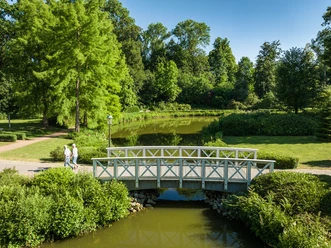 Preußisch Oldendorf-Kurensemble Bad Holzhausen-Teutoburger-Wald-Tourismus-D-Ketz-100.jpg Fußgängerbrücke über einen grünen Teich in einem üppigen Park mit Bäumen und Spaziergängern.