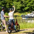 Preußisch Oldendorf-Mittellandkanal-Teutoburger-Wald-Tourismus-D-Ketz-084.jpg Zwei Radfahrer am Flussufer, winken einem vorbeifahrenden Boot, umgeben von grüner Natur.