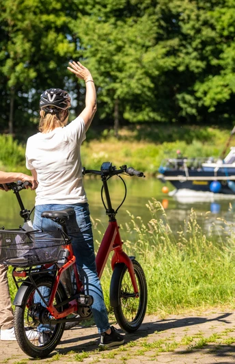 Preußisch Oldendorf-Mittellandkanal-Teutoburger-Wald-Tourismus-D-Ketz-084.jpg Zwei Radfahrer am Flussufer, winken einem vorbeifahrenden Boot, umgeben von grüner Natur.