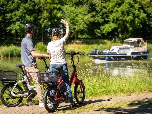 Preußisch Oldendorf-Mittellandkanal-Teutoburger-Wald-Tourismus-D-Ketz-084.jpg Zwei Radfahrer am Flussufer, winken einem vorbeifahrenden Boot, umgeben von grüner Natur.