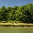 Preußisch Oldendorf-Mittellandkanal-Teutoburger-Wald-Tourismus-D-Ketz-090.jpg Zwei Radfahrer fahren entlang eines grünen Flussufers unter klarem, blauem Himmel in Preußisch Oldendorf.