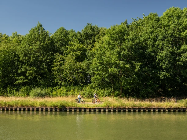 Preußisch Oldendorf-Mittellandkanal-Teutoburger-Wald-Tourismus-D-Ketz-090.jpg Zwei Radfahrer fahren entlang eines grünen Flussufers unter klarem, blauem Himmel in Preußisch Oldendorf.