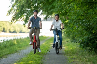 Preußisch Oldendorf-Mittellandkanal-Teutoburger-Wald-Tourismus-D-Ketz-097.jpg Zwei Menschen fahren lächelnd auf einem Fahrradweg entlang eines Flusses, umgeben von grünen Bäumen.