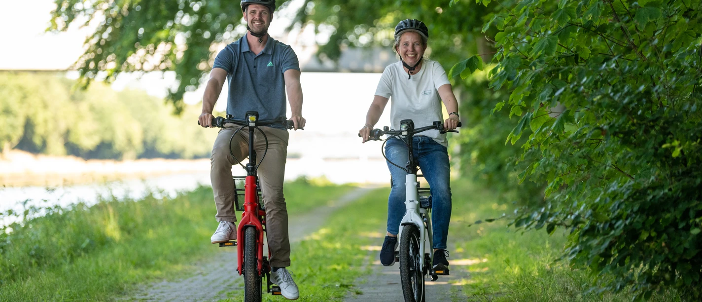 Preußisch Oldendorf-Mittellandkanal-Teutoburger-Wald-Tourismus-D-Ketz-097.jpg Zwei Menschen fahren lächelnd auf einem Fahrradweg entlang eines Flusses, umgeben von grünen Bäumen.