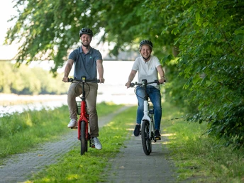 Preußisch Oldendorf-Mittellandkanal-Teutoburger-Wald-Tourismus-D-Ketz-097.jpg Zwei Menschen fahren lächelnd auf einem Fahrradweg entlang eines Flusses, umgeben von grünen Bäumen.