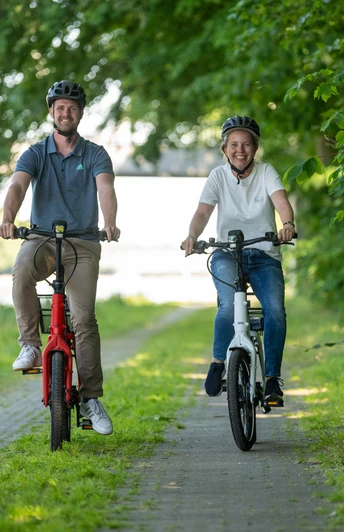 Preußisch Oldendorf-Mittellandkanal-Teutoburger-Wald-Tourismus-D-Ketz-097.jpg Zwei Menschen fahren lächelnd auf einem Fahrradweg entlang eines Flusses, umgeben von grünen Bäumen.