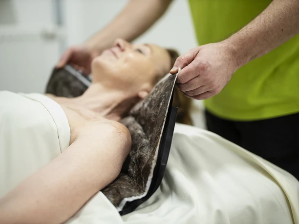 Moorpackung MEDICLIN Rose Klinik Eine Patientin erhält eine Moorpackung, betreut von einem Mitarbeiter in einem grünen Hemd.