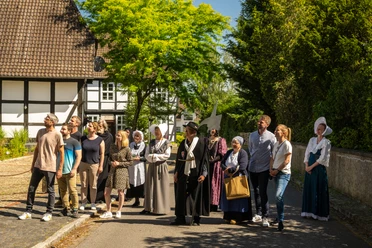 Stemwede-Stiftsort Levern-Teutoburger-Wald-Tourismus-D-Ketz-051.jpg Eine Gruppe Menschen in historischer Kleidung steht in einem sonnigen Dorf vor Fachwerkhäusern.