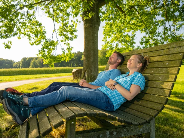 Stemwede-Waldlehrpfad Stemweder Berg-Teutoburger-Wald-Tourismus-D-Ketz-028.jpg Zwei Personen entspannen auf einer Holzbank unter einem Baum in sonniger, grüner Naturlandschaft.