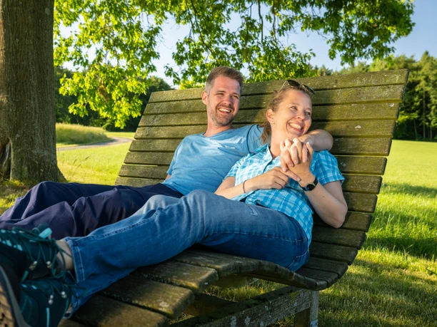 Stemwede-Waldlehrpfad Stemweder Berg-Teutoburger-Wald-Tourismus-D-Ketz-031.jpg Ein Paar genießt sonnige Stunden auf einer großen Holzbank im Grünen des Stemwede-Waldlehrpfads.