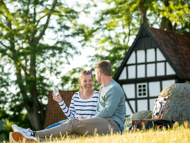 Paar sitzt auf Wiese vor Fachwerkhaus; sonniger Tag mit Bäumen im Hintergrund, entspanntes Picknick.