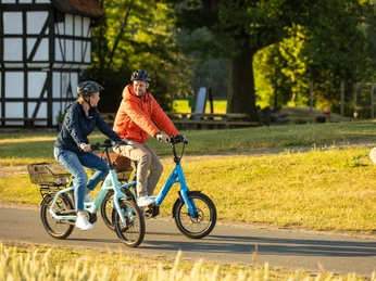 Stemwede-Mühlenensemble Levern-Teutoburger-Wald-Tourismus-D-Ketz-023.jpg Zwei Personen fahren an einem Fachwerkhaus in einer grünen Umgebung in Stemwede auf Fahrrädern.