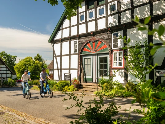 Stemwede-Stiftsort Levern-Teutoburger-Wald-Tourismus-D-Ketz-043.jpg Fachwerkhaus in Levern umgeben von grüner Landschaft, zwei Radfahrer auf ruhiger Straße davor.