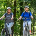 SHS-RadwegR1-Teutoburger-Wald-Tourismus-Patrick-Gawandtka-066.jpg Zwei lächelnde Frauen in Sportkleidung radeln durch einen grünen Wald auf E-Bikes.
