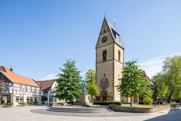 Steinhagen-Kirchplatz-Teutoburger-Wald-Tourismus-Patrick-Gawandtka-046-CC-BY-SA.jpg Historische Kirche mit Turmuhr und Fachwerkhäusern daneben. Zentraler Platz mit Statue und Bäumen.