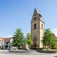 Steinhagen-Kirchplatz-Teutoburger-Wald-Tourismus-Patrick-Gawandtka-046-CC-BY-SA.jpg Historische Kirche mit Turmuhr und Fachwerkhäusern daneben. Zentraler Platz mit Statue und Bäumen.