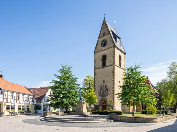 Steinhagen-Kirchplatz-Teutoburger-Wald-Tourismus-Patrick-Gawandtka-046-CC-BY-SA.jpg Historische Kirche mit Turmuhr und Fachwerkhäusern daneben. Zentraler Platz mit Statue und Bäumen.