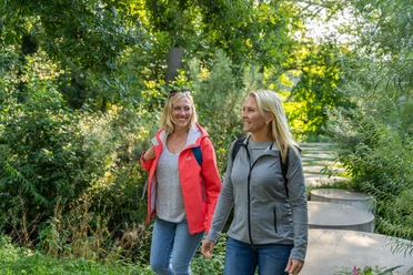 Steinhagen-Buergerpark-Teutoburger-Wald-Tourismus-Patrick-Gawandtka-016.jpg Zwei Frauen wandern lächelnd auf einem grünen Waldweg, umgeben von sommerlicher Vegetation.