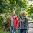 Steinhagen-Buergerpark-Teutoburger-Wald-Tourismus-Patrick-Gawandtka-016.jpg Zwei Frauen wandern lächelnd auf einem grünen Waldweg, umgeben von sommerlicher Vegetation.