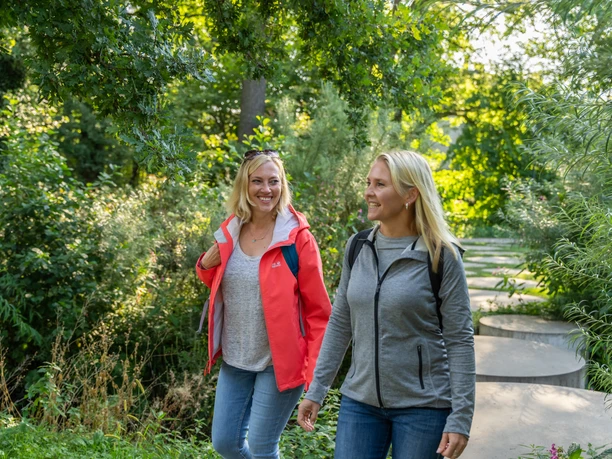 Steinhagen-Buergerpark-Teutoburger-Wald-Tourismus-Patrick-Gawandtka-016.jpg Zwei Frauen wandern lächelnd auf einem grünen Waldweg, umgeben von sommerlicher Vegetation.