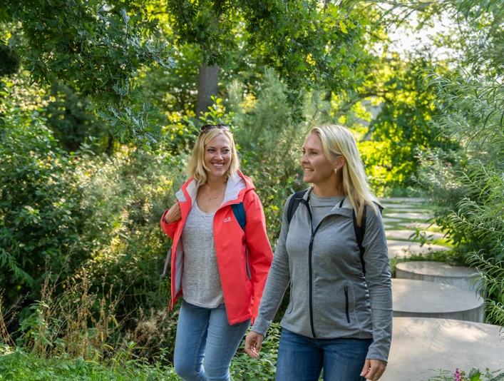 Steinhagen-Buergerpark-Teutoburger-Wald-Tourismus-Patrick-Gawandtka-016.jpg Zwei Frauen wandern lächelnd auf einem grünen Waldweg, umgeben von sommerlicher Vegetation.