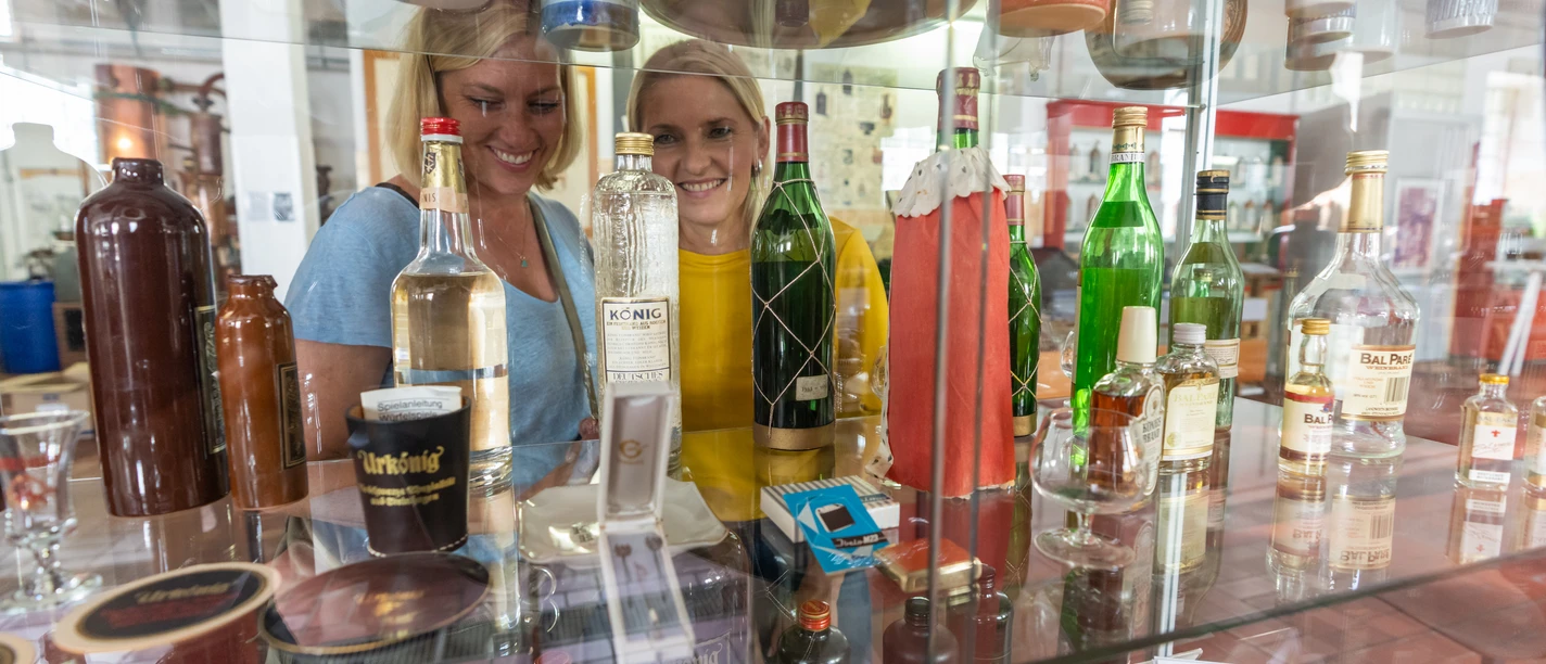 Steinhagen-HistorischesMuseum-Teutoburger-Wald-Tourismus-Patrick-Gawandtka-036.jpg Zwei Frauen betrachten Flaschen in einem Regal, umgeben von verschiedenen Spirituosen in einer Ausstellung.