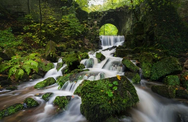 Een kleine waterval stroomt over met mos bedekte stenen onder een oude stenen brug in een groen, bebost gebied.