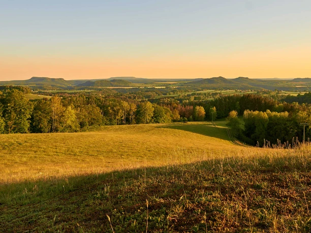 Gossdorf Hankehübel View Vast landscape with rolling hills and forests in the golden light of the sunset, the sky is clear and slightly orange in color.
