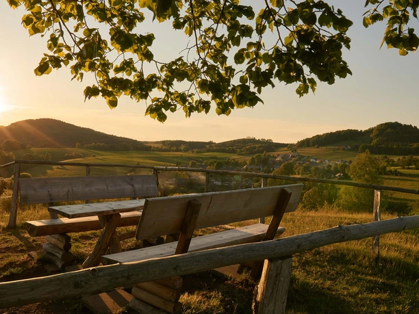 Goßdorf Hankehübel Uitzicht Houten banken en tafel op een heuvel met uitzicht op een groen landschap bij zonsondergang, omlijst door boomtakken.
