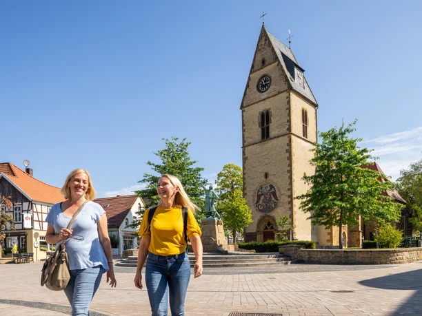 Steinhagen-Kirchplatz-Teutoburger-Wald-Tourismus-Patrick-Gawandtka-049.jpg Zwei lächelnde Frauen spazieren auf einem sonnigen Platz, im Hintergrund eine Kirche mit Uhrturm.