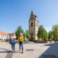 Zwei Frauen spazieren auf einem sonnigen Platz in Telgte, vorbei an einer Kirche mit Uhrturm.