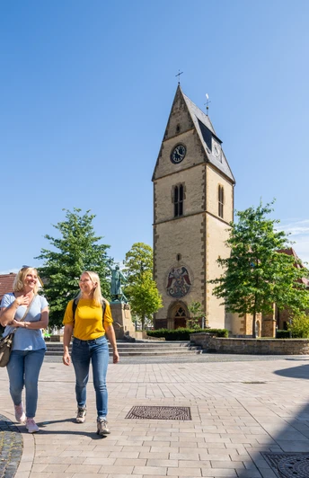 Zwei Frauen spazieren auf einem sonnigen Platz in Telgte, vorbei an einer Kirche mit Uhrturm.