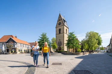 Steinhagen-Kirchplatz-Teutoburger-Wald-Tourismus-Patrick-Gawandtka-051.jpg Zwei Frauen spazieren auf einem sonnigen Platz in Telgte, vorbei an einer Kirche mit Uhrturm.