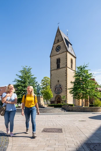Steinhagen-Kirchplatz-Teutoburger-Wald-Tourismus-Patrick-Gawandtka-051.jpg Zwei Frauen spazieren auf einem sonnigen Platz in Telgte, vorbei an einer Kirche mit Uhrturm.