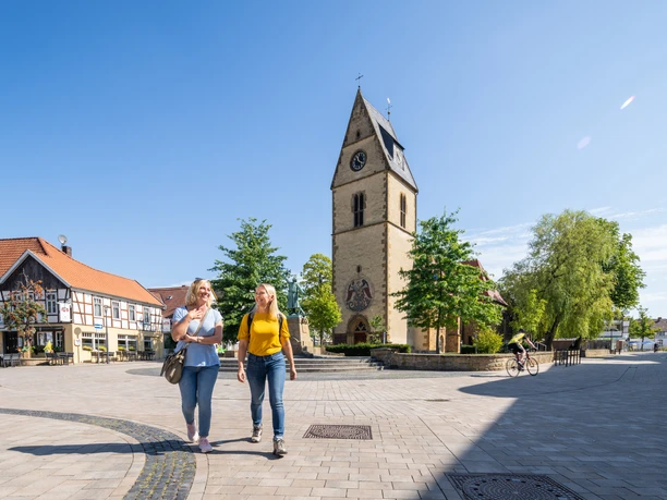 Steinhagen-Kirchplatz-Teutoburger-Wald-Tourismus-Patrick-Gawandtka-051.jpg Zwei Frauen spazieren auf einem sonnigen Platz in Telgte, vorbei an einer Kirche mit Uhrturm.