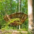 Zwei Personen betrachten eine große hölzerne Hängeskulptur im Wald bei sonnigem Wetter.