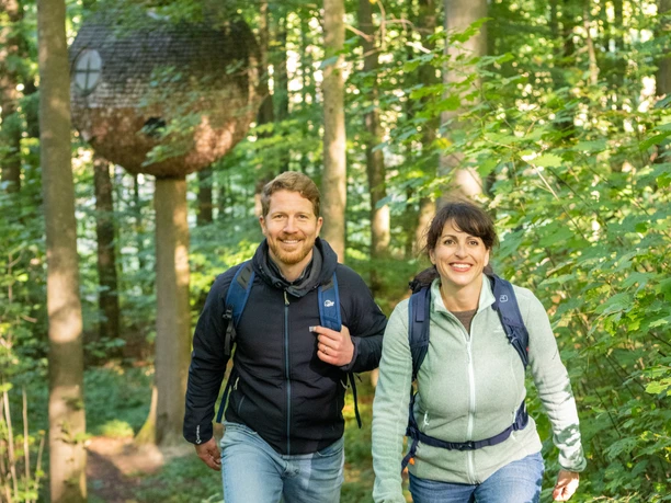 Nieheim-Nieheimer Kunstpfad Holzhausen-Teutoburger-Wald-Tourismus-D-Ketz-031.jpg Paar wandert freudig durch einen grünen Wald mit einem baumelnden kugelförmigen Baumhaus im Hintergrund.