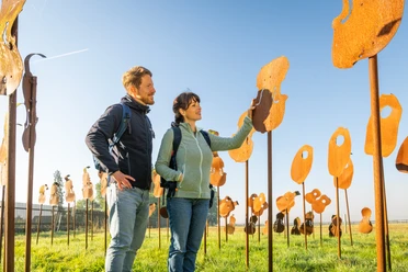 Nieheim-Nieheimer Kunstpfad Holzhausen-Teutoburger-Wald-Tourismus-D-Ketz-003.jpg Mann und Frau stehen auf einer Wiese, umgeben von Metallskulpturen, bei klarem Himmel.
