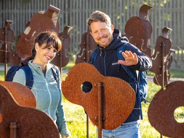 Nieheim-Nieheimer Kunstpfad Holzhausen-Teutoburger-Wald-Tourismus-D-Ketz-004.jpg Zwei lächelnde Personen interagieren mit rostigen Gitarren-Skulpturen auf einer sonnigen Wiese.