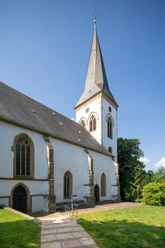 Oerlinghausen-Alexanderkirche-Teutoburger-Wald-Tourismus-Patrick-Gawandtka-057-CC-BY-SA.jpg Eine weiße Kirche mit spitzem Turm unter wolkenlosem Himmel in grüner, ländlicher Umgebung.