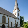 Oerlinghausen-Alexanderkirche-Teutoburger-Wald-Tourismus-Patrick-Gawandtka-057-CC-BY-SA.jpg Eine weiße Kirche mit spitzem Turm unter wolkenlosem Himmel in grüner, ländlicher Umgebung.