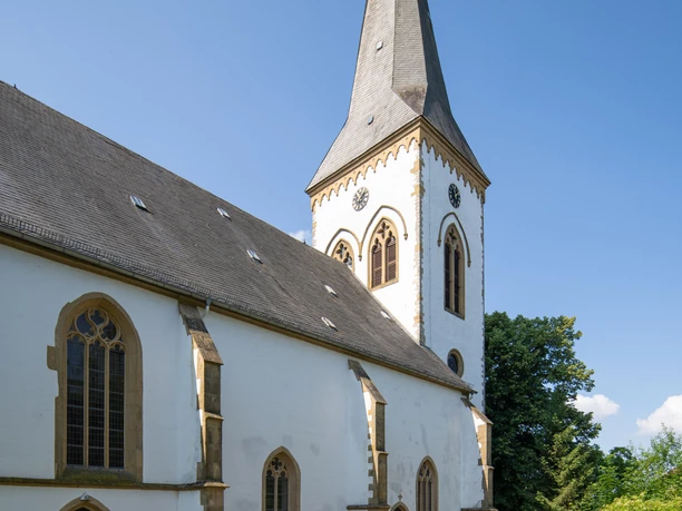 Oerlinghausen-Alexanderkirche-Teutoburger-Wald-Tourismus-Patrick-Gawandtka-057-CC-BY-SA.jpg Eine weiße Kirche mit spitzem Turm unter wolkenlosem Himmel in grüner, ländlicher Umgebung.