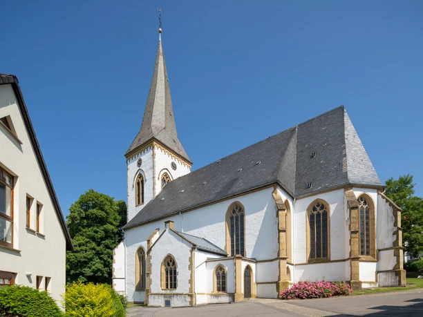 Klassische Kirche mit hohem Turm und Spitzdach, umgeben von grünen Bäumen unter klarem Himmel.