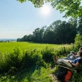 Zwei Personen entspannen auf einer Holzbank am Waldrand mit Blick auf eine weite, sonnige Wiese.