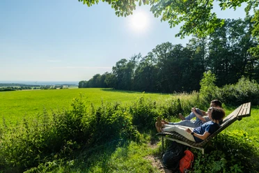 Schlangen-Bauerkamp-Teutoburger-Wald-Tourismus-Patrick-Gawandtka-098.jpg Zwei Personen entspannen auf einer Holzbank am Waldrand mit Blick auf eine weite, sonnige Wiese.