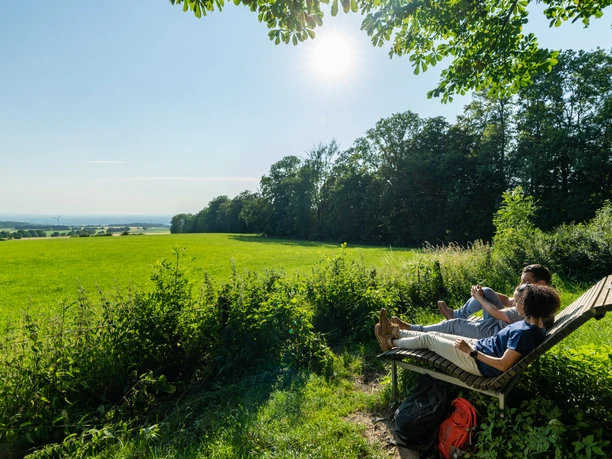 Schlangen-Bauerkamp-Teutoburger-Wald-Tourismus-Patrick-Gawandtka-098.jpg Zwei Personen entspannen auf einer Holzbank am Waldrand mit Blick auf eine weite, sonnige Wiese.