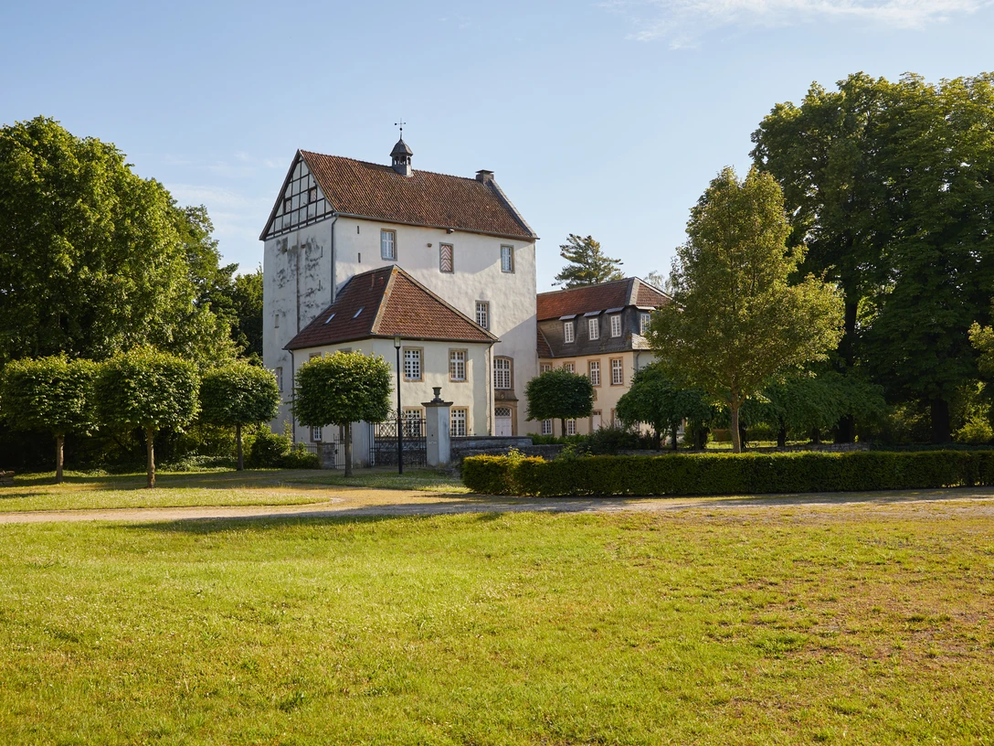 Salzkotten-Burg-Dreckburg-Teutoburger-Wald-Tourismus-T-Evers-003-CC-BY-SA.jpg Ein historisches Herrenhaus mit Fachwerk, umgeben von gepflegtem Rasen und grünen Bäumen im Sonnenschein.